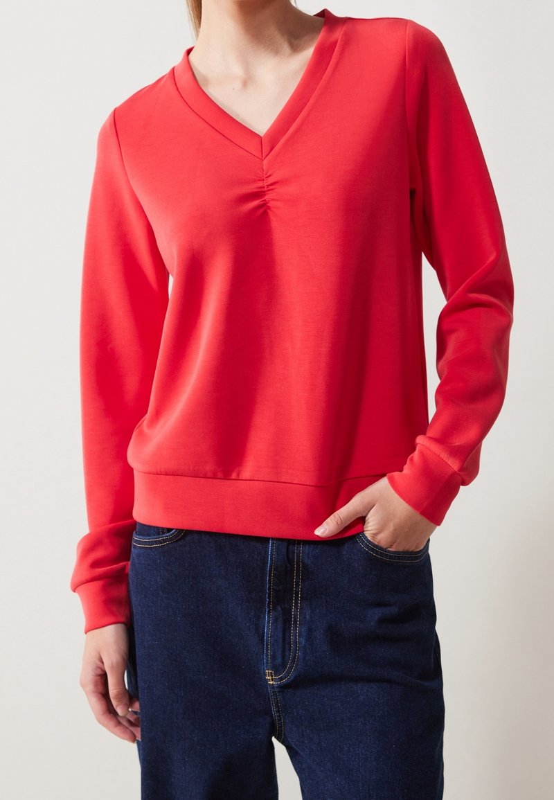 Suéter rojo de manga larga con escote en V y detalle fruncido en el cuello. Tejido suave y puños acanalados. Combinado con jeans de mezclilla oscura.