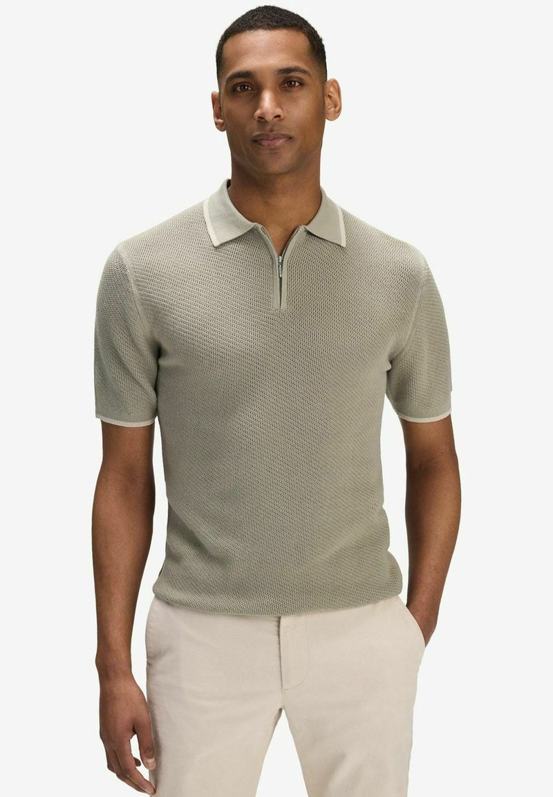 Homme portant un polo en maille texturée gris clair avec un col zippé et un pantalon beige, debout avec une main dans la poche.
