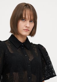 Blusa nera in pizzo con un design aderente, abbottonatura frontale, maniche corte a sbuffo e vari motivi texture in tutto il capo.
