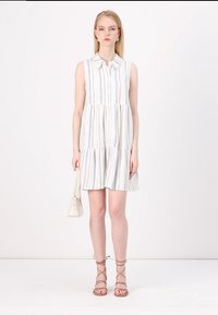 Robes blanches sans manches avec des rayures horizontales bleues, design à niveaux et col. La mannequin tient un petit sac beige et porte des sandales à lanières marron clair.
