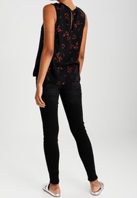 Svart blommig blus med en nyckelhålsdetalj bak, ihopparad med slim fit svarta jeans. Modell som bär platta silverfärgade sandaler.