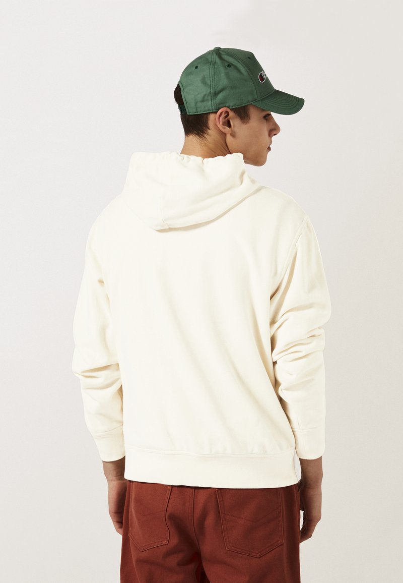 Haut à capuche beige avec cordon de serrage et poche kangourou, porté avec un pantalon couleur rouille. Accompagné d'une casquette verte avec un logo.