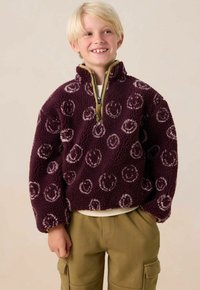 Fleece pullover in diep paars met een crèmekleurig smiley-achtig patroon, kwart rits en groene kraag. Gecombineerd met olijfgroene cargo broeken.