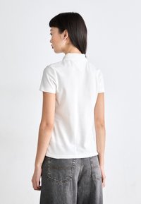 Polo shirt blanc en coton côtelé, à manches courtes, col classique, associé à un short en denim gris taille haute avec des poches visibles.