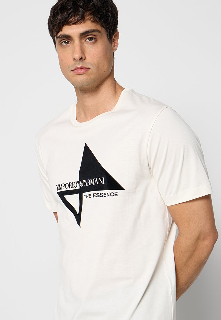 Joven con camiseta blanca Emporio Armani con logo geométrico negro y texto "The Essence" sobre fondo liso claro.