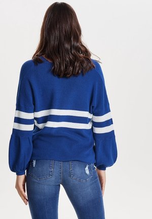 Vrouw met lang bruin haar, gekleed in een blauwe trui met witte strepen en opgeblazen mouwen, gecombineerd met versleten blauwe spijkerbroeken, kijkt weg van de camera.