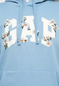 Light blue hoodie with white GAP letters adorned by white and orange floral designs, featuring a front pocket and drawstrings.