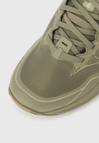 Chaussure de sport vert olive avec une tige en mesh texturé, un embout renforcé et un système de laçage. Détail de marque latérale notable.