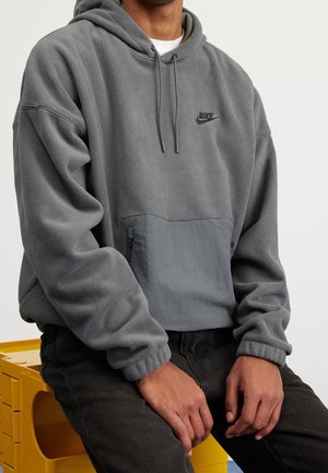 Person in einem grauen Nike-Fleece-Hoodie und schwarzen Hosen sitzt auf einer gelben Kiste, die Hände liegen auf den Knien, neutraler Hintergrund.