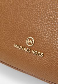Lähikuva tekstuuripinnasta ruskeasta nahasta, jossa on kultainen Michael Kors -logo ja pyöreä MK-symboli käsilaukussa tai asusteessa.