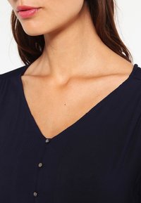 Femme portant un haut bleu marine à col en V avec de petits boutons ronds sur le devant, laissant apparaître son cou et la zone de sa clavicule.