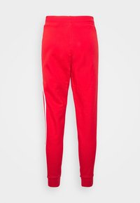 Červené tepláky s užším střihem, elastickým pasem a bílými postranními pruhy. Vyrobené z měkké, hladké látky s žebrovanými lemy u kotníků.