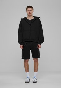Melnas zip-up kapuces jaka ar kangaroo kabatu, apvienota ar melniem džinsu šortiem ar nodilušiem akcentiem un ķēdes detaļu. Klasiskie sporta apavi un baltas zeķes.