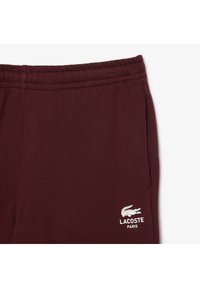 Burgundy sweatpants med en slät textur, med en vit broderad Lacoste-logotyp och elastisk midja. En sidoficka synlig.