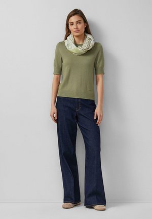 Femme debout devant un fond uni, portant un haut à manches courtes vert olive, une écharpe vert clair à motifs, un jean bleu foncé et des chaussures beiges.