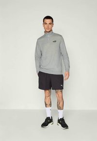 Šedá mikina na zip s černým logem, černé šortky a černé sportovní boty. Model stojí před neutrálním pozadím.