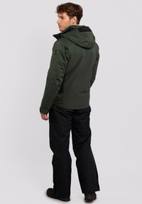 Giacca isolata verde scuro con cappuccio, caratterizzata da un pannello posteriore nero e un logo, indossata con pantaloni neri con zip alle caviglie.