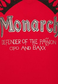 Rotes Baumwoll-T-Shirt mit großem grauem Grafiktext "Monarch" und silbernen Akzenten. Der Text enthält außerdem "DEFENDER OF THE PASSION" und "CIPO AND BAXX."