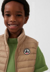 Beige gewatteerd vest met hoge kraag en een logopatch. Gedragen over een groen T-shirt, wat een casual gelaagde outfit laat zien.