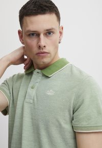 Grønn polo skjorte med teksturert stoff, hvit krage og kremfargede detaljer. Har et lite brodert logo på brystet.