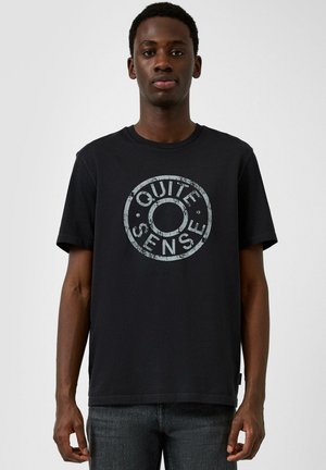 Mężczyzna w czarnej koszulce z wyblakłym okrągłym logo "QUITE SENSE", stojący na tle jednolitego jasnoszarego tła.