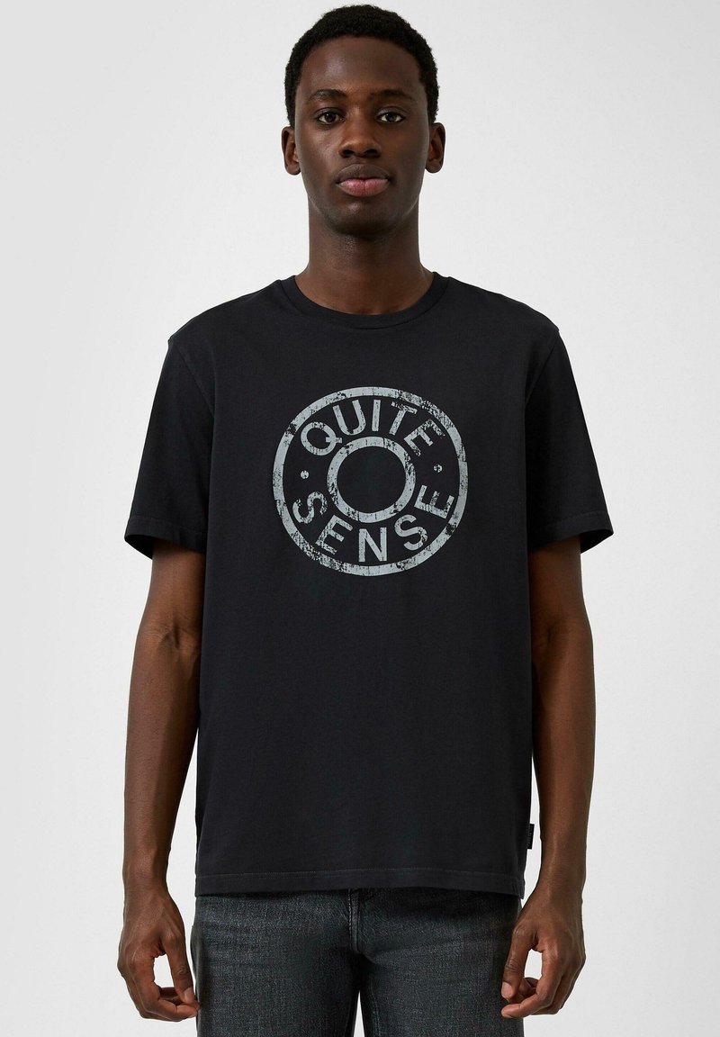 Muž v černém tričku s vybledlým kulatým logem "QUITE SENSE", stojící před jednoduchým světle šedým pozadím.