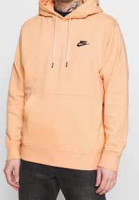 Persikofärgad bomullshoodie med ficka framtill, justerbara snören, svart Nike-logga på bröstet, samt ribbade manschetter och fåll.