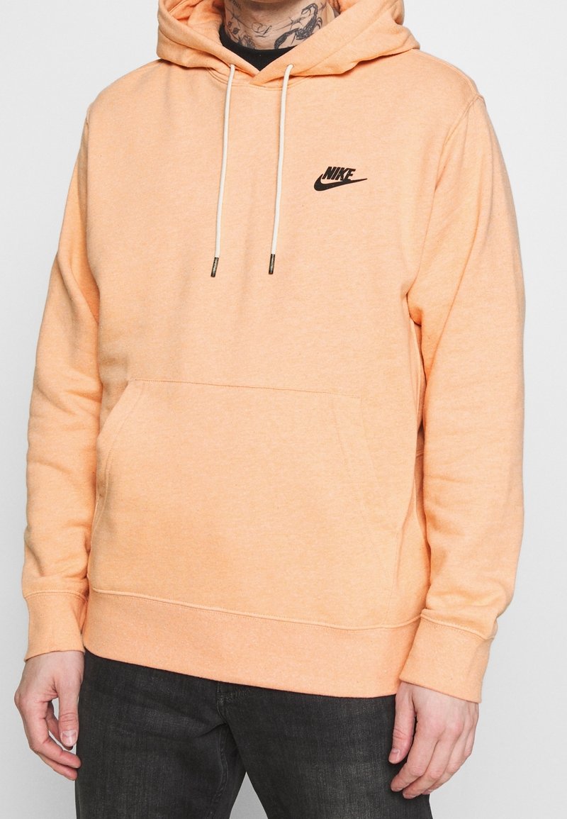 Persikofärgad bomullshoodie med ficka framtill, justerbara snören, svart Nike-logga på bröstet, samt ribbade manschetter och fåll.