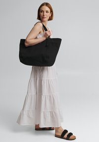 Zwarte tote bag met een gestructureerd ontwerp, voorzien van een brede opening, stevige handvatten en een gladde canvas textuur. Model draagt een witte gelaagde jurk.