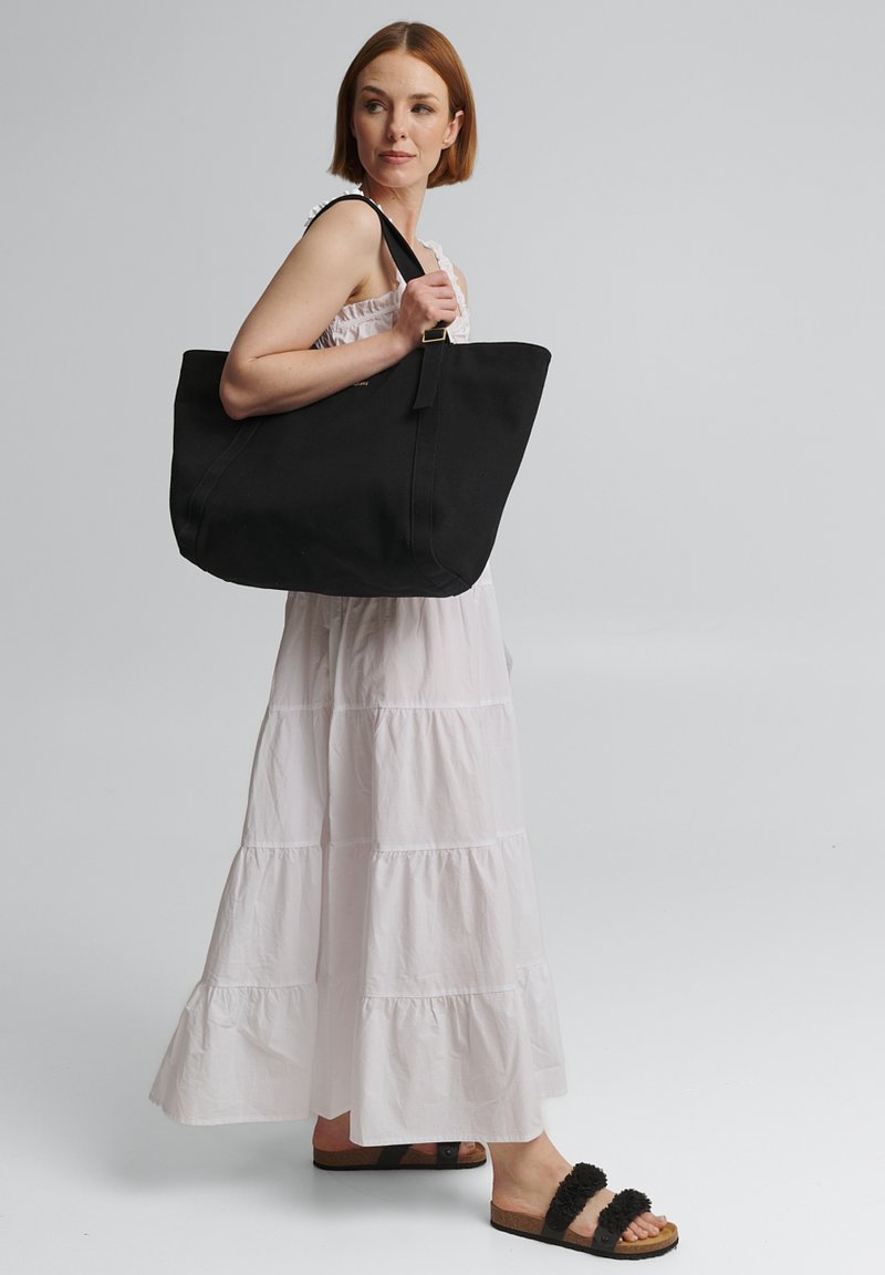 Zwarte tote bag met een gestructureerd ontwerp, voorzien van een brede opening, stevige handvatten en een gladde canvas textuur. Model draagt een witte gelaagde jurk.