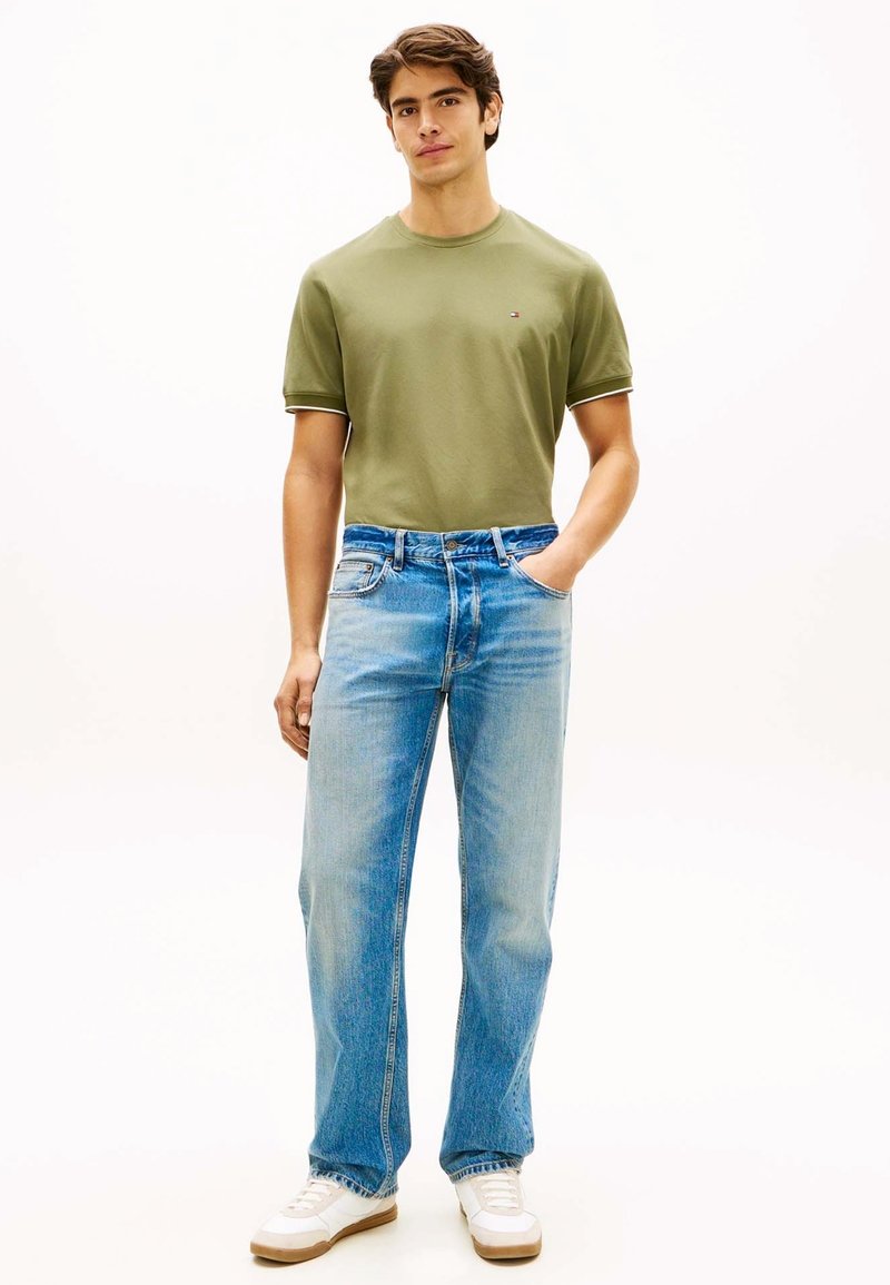 Jeune homme debout avec une main dans la poche, portant un t-shirt vert olive à manches courtes, un jean bleu clair et des baskets blanches sur fond blanc.