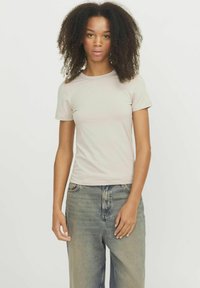 T-shirt aderente beige chiaro con maniche corte, realizzato in tessuto morbido, abbinato a jeans blu larghi, che mostrano una silhouette rilassata.