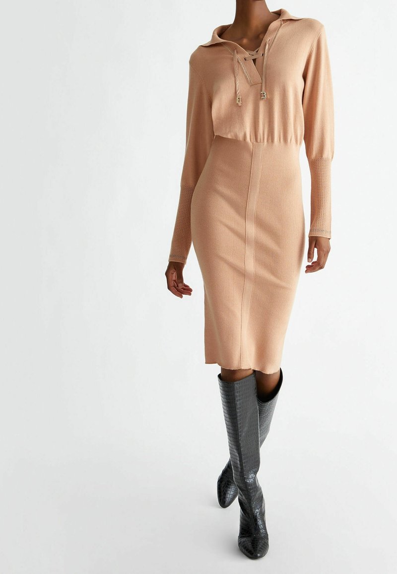 Femme portant une robe en maille beige ajustée à manches longues et col, associée à des bottes noires texturées montant jusqu'aux genoux, debout sur un fond blanc.