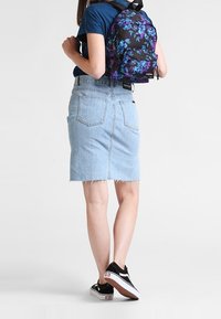 Zwarte rugzak met paarse en blauwe bloemenpatroon, gedragen op de rug van een persoon, gecombineerd met een lichte spijkerrok en zwarte sneakers.