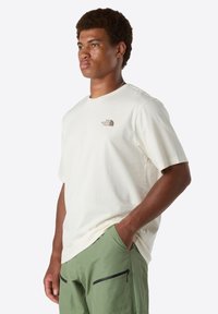 Jeune homme portant un t-shirt beige The North Face et un pantalon vert avec poches zippées, se tenant avec une main dans sa poche.