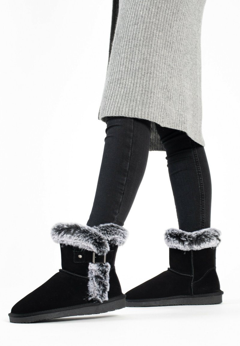 Botines de ante negro con borde de pelo sintético, detalle de hebilla y suelas de goma planas. Usados con jeans negros y un suéter de punto gris.