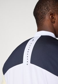 Koszula sportowa z kołnierzykiem z kolorowym podziałem w odcieniach granatowym, białym i kremowym. Wyraziste logo w czerni na plecach.