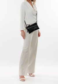 Borsa a spalla in pelle nera con una tasca con zip, abbinata a un top drappeggiato bianco e pantaloni palazzo beige. Sandali semplici con cinghie.