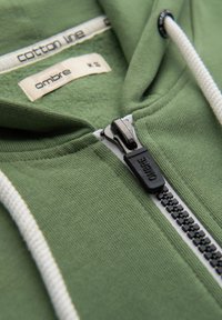 Sweat à capuche vert en coton avec cordons blancs, fermeture éclair visible étiquetée "OMBRE" et étiquettes de vêtements indiquant "cotton line" et "ombre M 36".