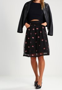 Žena v černém krátkém svetru, černé květinové sukni s červenými květy, černé kožené bundě přehozené přes ramena a černých lakovaných lodičkách.