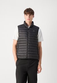 Schwarze gepolsterte Weste mit hohem Kragen, Frontreißverschluss und Seitentaschen. Über einem hellgrauen Shirt getragen, das eine glatte, glänzende Textur zeigt.