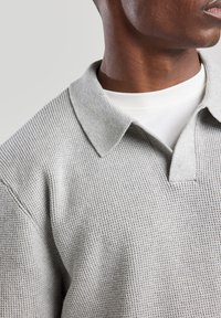 Harmaa neulottu polo-pusero, jossa on lyhyet kaulukset ja teksturoitu kuvio, valkoisen pyöreäkauluksisen paidan päällä, rento istuvuus.