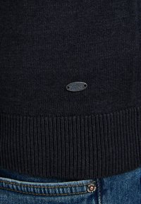 Dunkelblauer, gestrickter Pullover mit geripptem Saum, ausgestattet mit einem kleinen ovalen Metalletikett. Kombiniert mit hellblauen Jeans in der Taille.
