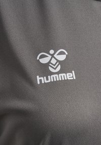 Szara tkanina o gładkiej fakturze z białym logo przedstawiającym abstrakcyjne kształty skrzydeł nad słowem „hummel”.