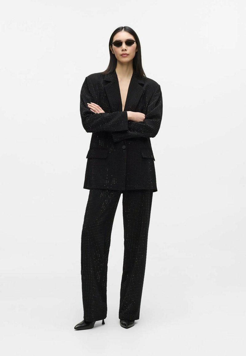 Femme portant un costume noir clouté, des talons noirs pointus et de petites lunettes de soleil noires, debout les bras croisés sur fond blanc.