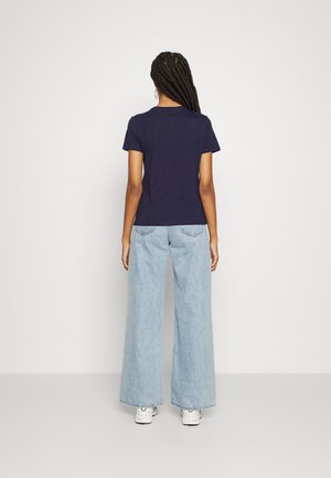 Mujer con cabello trenzado, vistiendo una camiseta de manga corta azul marino, jeans de pierna ancha color azul claro y zapatillas blancas, de pie de espaldas a la cámara.