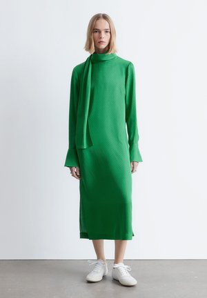 Groene jurk met lange mouwen, een hoge hals en een sjaaldetail; voorzien van kleine zwarte stippen, een losse pasvorm en zijsplitten. Gecombineerd met witte sneakers.