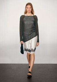 Schwarzes, offenes Strickoberteil mit langen Ärmeln und asymmetrischem Saum, kombiniert mit einem weißen Rock mit schwarzem Spitzenbesatz, accessorized mit einer schwarzen Clutch.