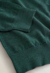 Tuvumā tumši zaļa adīta džemperu piedurkne ar ribotu aproci un salocītu audumu uz gaišas virsmas.