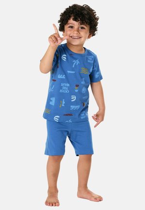 Lächelndes, lockiges Kind in blauen Shorts und bedrucktem T-Shirt, das mit den Fingern ein "L"-Zeichen macht, barfuß auf weißem Hintergrund stehend.